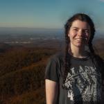 Women in 20's, Caucasion, with long dark brown hair in two braids. Wearing grey t-shirt with white graphic, background of mountain landscape and blue sky. My areas of interest include sustainable networking and community engagement, sustainable art, and the circular economy.